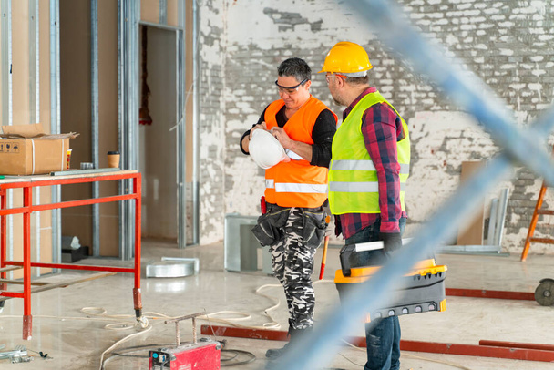 Construtor em uniforme de trabalho com capacete de proteção em pé com instrumentos no local de construção dentro de casa - Foto, Imagem