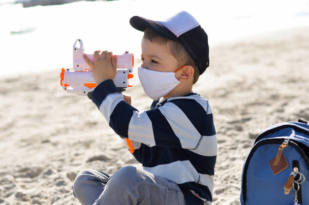 A cute four-year-old preschooler boy in a protective cotton mask sits on the beach and plays with his gun. - Photo, Image