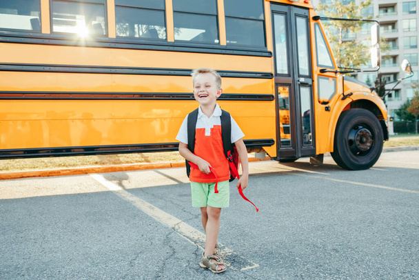 Gelukkige glimlachende blanke jongen elementaire student met rugzak in de buurt van de gele bus op de eerste september dag. Onderwijs en terug naar school in het najaar. Kind op school buiten op school.  - Foto, afbeelding