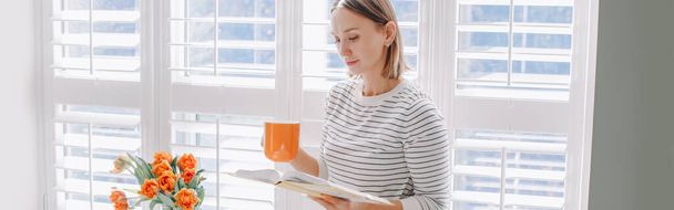 家で本を読んでいる美しい幸せな中年の女性。短髪の若い女性はマグカップからティーコーヒーを飲み、本を持っています。リラックスして、自宅で余暇時間。Webバナーヘッダー. - 写真・画像