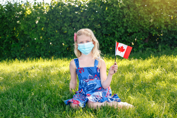 カナダコロナウイルスパンデミックでの休日のお祝い。顔のマスクを振ってカナダのフラグを保持の子の女の子。公園で草の上に座っている保護マスクの子供カナダの日を屋外で祝う. - 写真・画像