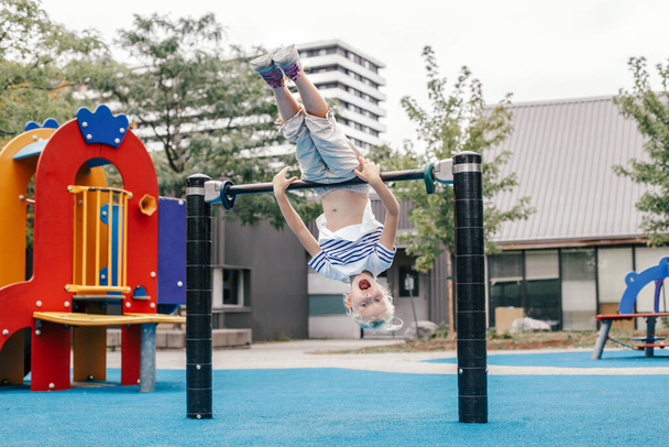 Happy smiling young girl exercising on children pull-ups bar outdoor at playground park. Child hanging on high bar upside down. Summer fun activity workout and fitness. Kids sports and recreation. - Photo, Image