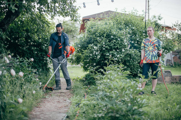 Δύο άντρες δουλεύουν στον κήπο. Κουρεύουν το γρασίδι με μια χειροκίνητη μηχανή και ποτίζουν θάμνους και λουλούδια. Συντήρηση κήπων και γκαζόν σε εξοχικό σπίτι - Φωτογραφία, εικόνα