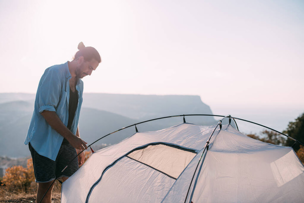 Giovane turista di sesso maschile mette una tenda in montagna. Il ragazzo allestisce un campo per un'escursione, raccoglie una tenda da solo in montagna al tramonto - Foto, immagini