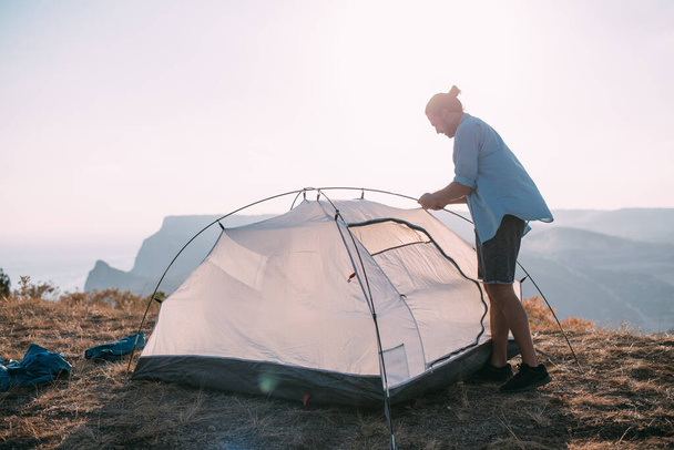 Молодой турист ставит палатку в горах. Парень разбивает лагерь в походе, собирает палатку в горах на закате - Фото, изображение