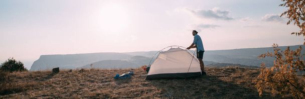 Young male tourist puts a tent in the mountains. The guy sets up a camp on a hike, collects a tent alone in the mountains at sunset - Photo, Image