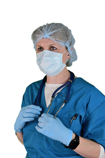 Female doctor in a blue medical suit, mask and cap. A stethoscope hangs around her neck, which she holds with both hands. Isolated on white background. - 写真・画像