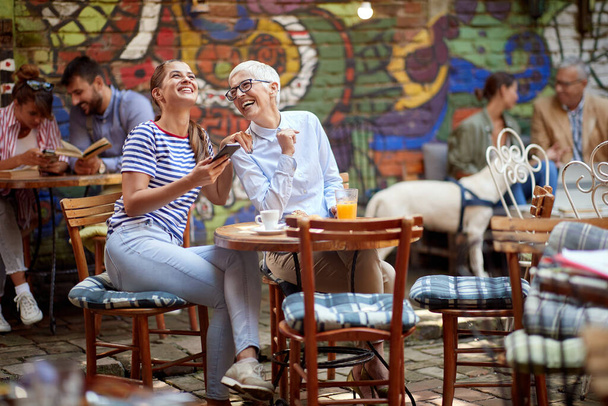 An older woman and her young female friend laughing about a smartphone content they watching while have a drink in a cheerful atmosphere in the bar. Leisure, bar, friendship, outdoor - Photo, image