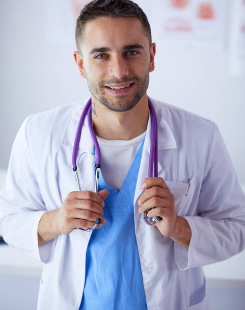 Young and confident male doctor portrait standing in medical office. - Фото, изображение