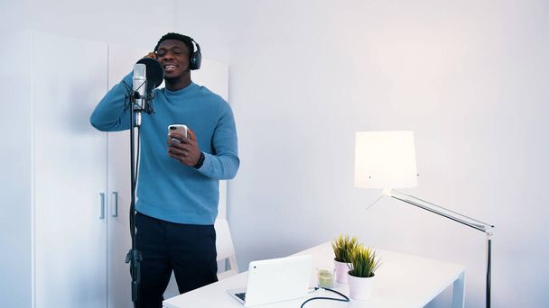 Hombre con auriculares cantando una canción en un micrófono en un estudio. Con un teléfono en la mano mirando la letra. - Foto, Imagen