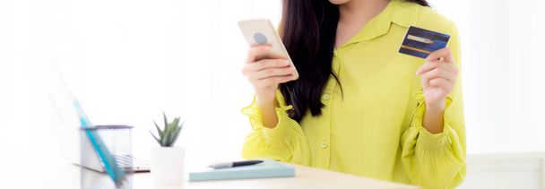 Молодая азиатская бизнес-женщина, использующая смартфон и держащая кредитную карту во время онлайн-покупок и оплаты онлайн с ноутбуком на столе дома, женщина держит дебетовую карту, коммуникационная концепция. - Фото, изображение