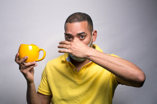 Латиноамериканец в маске сжимает нос, держа желтую чашку, изолированную на сером фоне - Фото, изображение