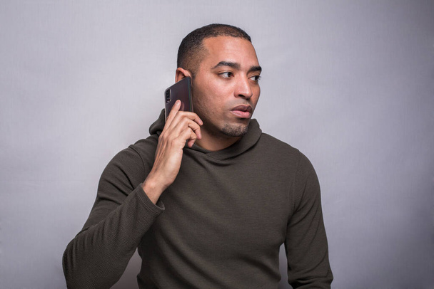 A portrait of a Hispanic, Latino Black man in green hoodie speaking on the phone, isolated on grey - Fotó, kép