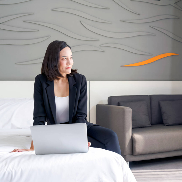 Una mujer asiática en traje negro usa computadora portátil para trabajar por la mañana en la cama. Miró afuera mientras pensaba en algo.. - Foto, imagen