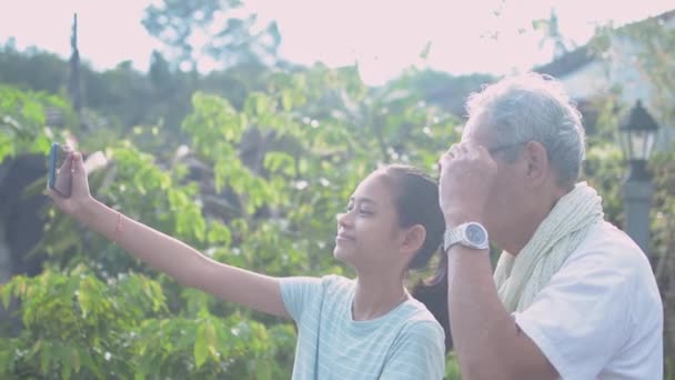 Ázsiai imádnivaló lány és az ő idős ősz haj nagyapja élvezik, hogy szelfi fotó együtt okostelefon a ház előtt. A családi kötelék. Egészséges életmód. - Felvétel, videó