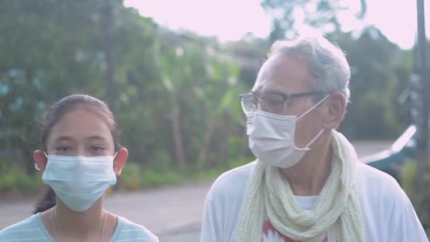 Imádnivaló thai lány, szemüveges idősebb nagyapjával, arcmaszkot visel, és kényelmesen sétál együtt a vidéki faluban. Minden nap maszk viselése. Egészséges életmód. - Felvétel, videó