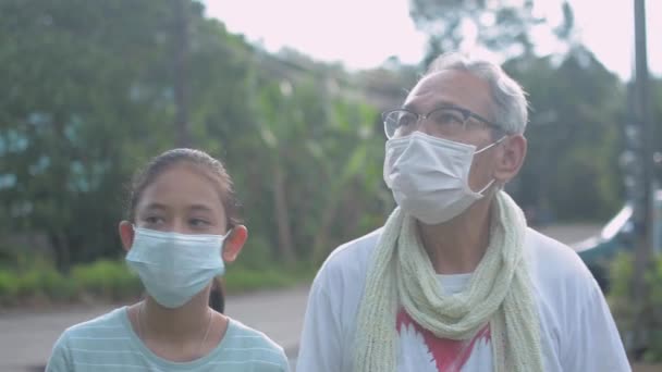 アジア系のシニア白髪の祖父は、顔のマスクを着て、午前中に農村部の村で彼の愛らしい若い孫娘とゆっくりと話をして歩く。幸福と健康的なライフスタイル.  - 映像、動画