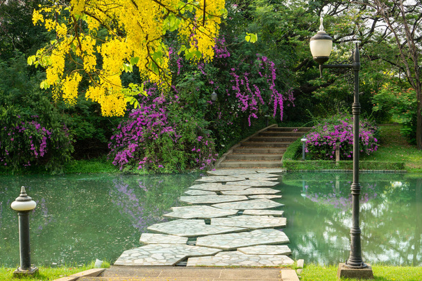 Ponte de concreto de curva de pedra de passo cinza e passarela cruzam um pequeno lago entre árvores verdes, flores e arbustos em um parque e boa manutenção de cuidados paisagísticos - Foto, Imagem
