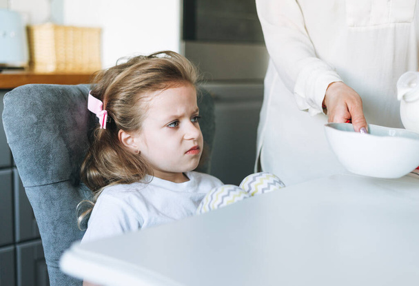 Ongelukkig klein kind mooi meisje met lang haar in huis kleding met yoghurt zitten aan eettafel in de keuken thuis. Het kind wil niet eten. - Foto, afbeelding