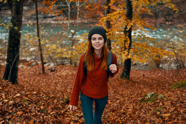 un viajero en un suéter rojo y un sombrero camina en el bosque de otoño cerca de una vista de la cima del río de montaña - Foto, Imagen