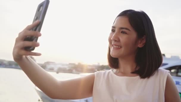 Happy young Asian vrouw blogger toerist met casual stijl gebruik smartphone nemen selfie genieten met mooie zonsondergang rivier uitzicht haven in cafe in de stad, Lifestyle toeristische reizen concept. - Video