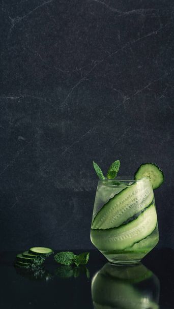 Uitzicht op drinkglas met komkommer tonic gin cocktail met verse munt op glanzende zwarte tafel en achtergrond verfrissende lange drank zomer recept - Foto, afbeelding