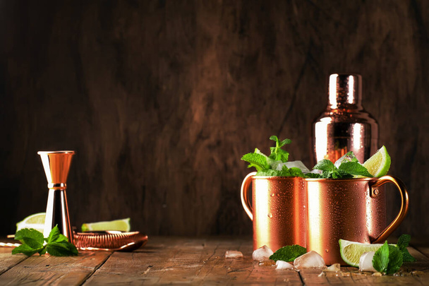 Moscow mule or mint julep alcoholic cocktail in copper mug with lime, ginger beer, vodka and mint. Wooden table, copper bar tools - Φωτογραφία, εικόνα