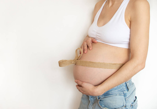 Embarazada vientre con lona ecológica cinta de lino alrededor del vientre, vista de perfil. Mujer embarazada joven en jeans sobre un fondo de pared. Lugar para el texto - Foto, imagen