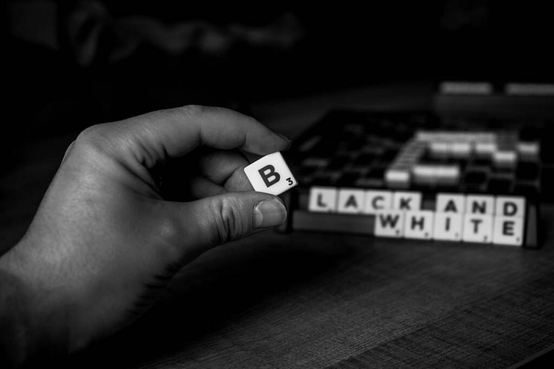 Brecht, Belçika - 28 Temmuz 2020: Bir kişinin siyah beyaz portresi üzerinde siyah ve beyaz yazılar olan Scrabble tahtası önünde parmaklarının arasında b harfini tutan bir kişinin.. - Fotoğraf, Görsel