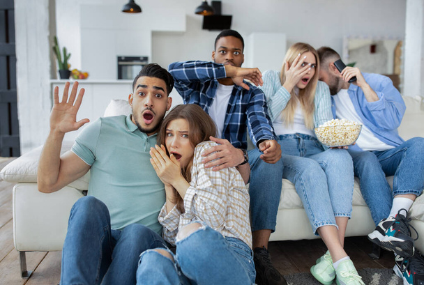 Frightened millennial friends watching scary movie or thriller, holding popcorn, covering their faces in horror at home - Foto, Bild