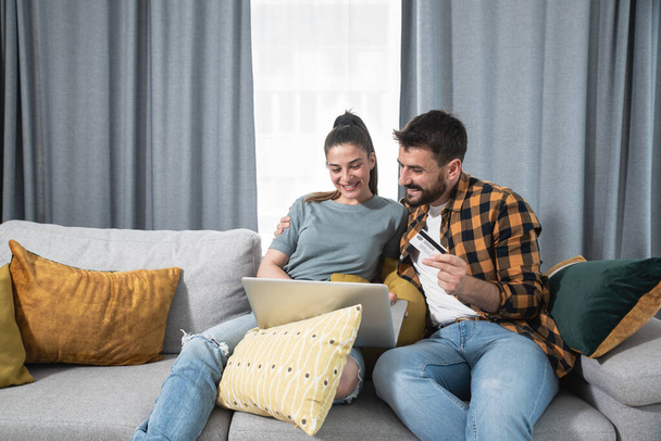 Young happy couple with laptop computer sitting at home ordering and online shopping a new furniture and kitchen equipment for their apartment cause they start living together  - Photo, Image
