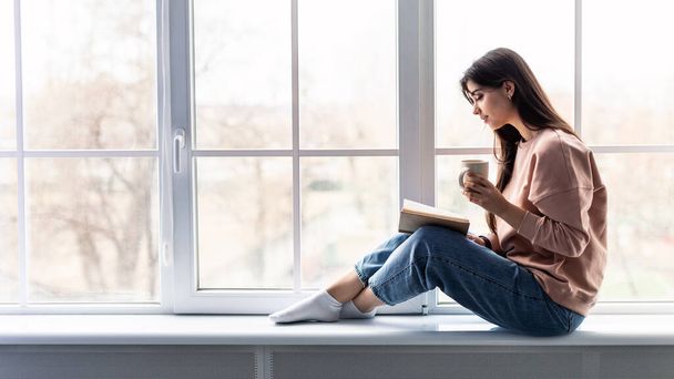 Vrouw zit op vensterbank koffie te drinken leesboek - Foto, afbeelding