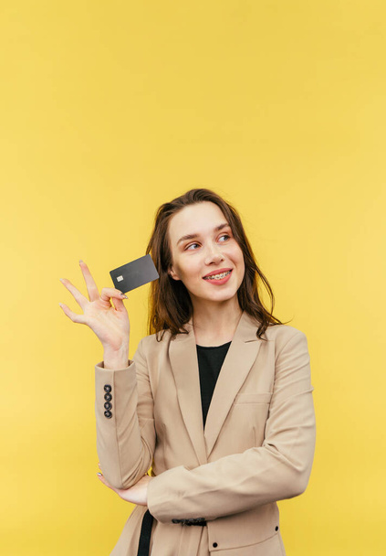Cute positive lady in a beige jacket poses with a bank card on a yellow background and looks away with a smile on her face. - 写真・画像