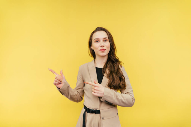 Positive lady in a beige jacket stands on a yellow background with a smile on her face looks at the camera and shows her fingers to the side on copy space - Photo, Image