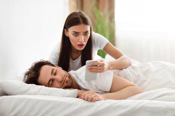 Geschokt vriendin lezen berichten op bedriegende echtgenoten telefoon in de slaapkamer - Foto, afbeelding