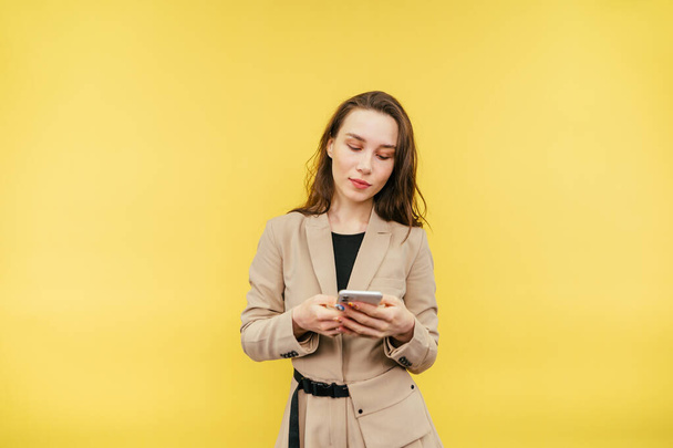 Gyönyörű nő okos ruhák használ egy okostelefon sárga alapon, nézi a képernyőn egy nyugodt arc, visel bézs kabát. - Fotó, kép