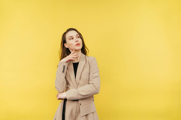 Portrait of a pensive female worker in a beige suit standing on a yellow background, looking away and thinking. Copy space - Zdjęcie, obraz