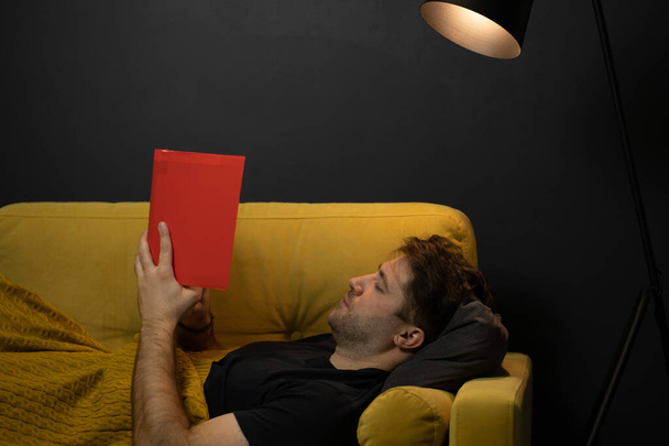 Young unshaven handsome guy reads book liyng on comfortable yellow sofa - Photo, Image
