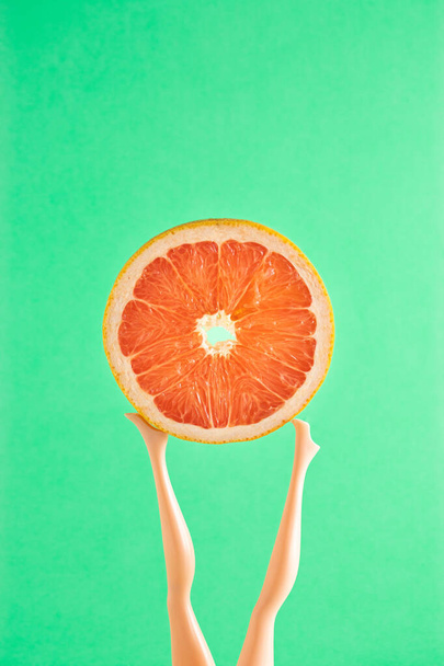 Summer beach creative idea. A doll holding a slice of fresh red grapefruit on its feet. Minimal fruit or healthy food concept. - Foto, Imagem