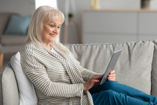 Smiling mature woman using tablet sitting on couch - Valokuva, kuva