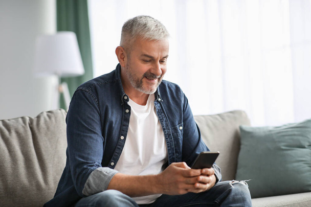 Smiling grey-haired mature man sitting on couch, using mobile phone - Φωτογραφία, εικόνα