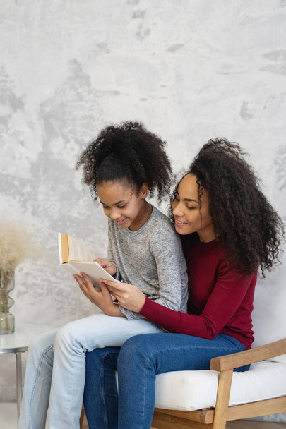 Mamá afroamericana con su hija leyendo un libro sentado en una silla en casa. - Foto, imagen