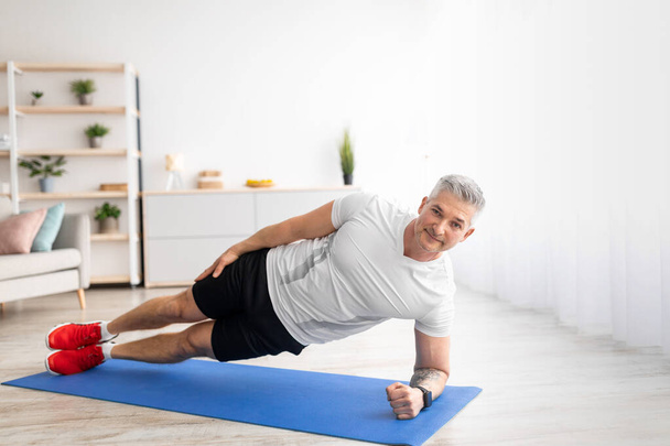 Gesunder Lebensstil. Fit reifer Mann steht zu Hause in Seitenplanke auf Sportmatte, trainiert im Wohnzimmer - Foto, Bild