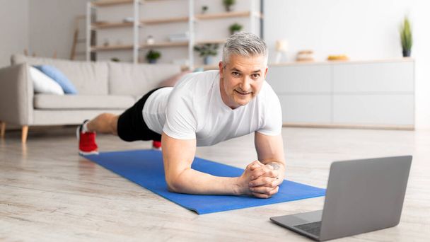 Sabah antrenmanı. Sportif orta yaşlı adam yoga tahtası yapıyor, dizüstü bilgisayarda online ders izliyor, oturma odasında egzersiz yapıyor. - Fotoğraf, Görsel