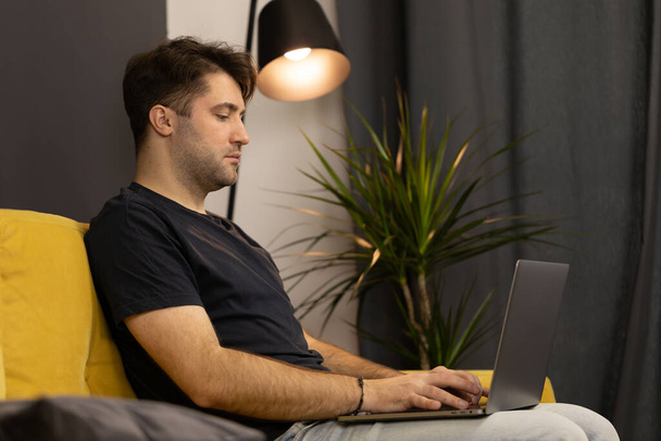 Genç, tıraşsız yakışıklı adam kanepede oturuyor ve dizüstü bilgisayarında klavye çalıyor. - Fotoğraf, Görsel