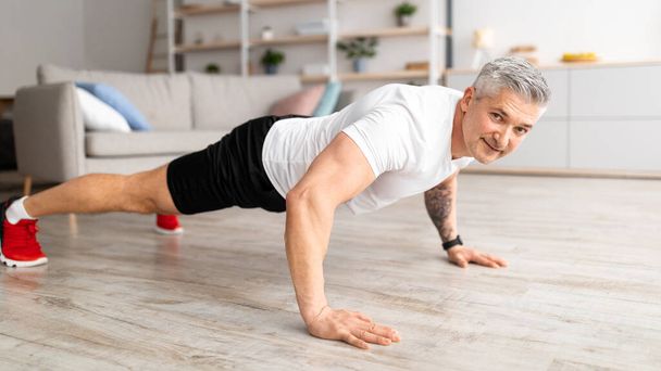 Mannelijke fitness training routine. Happy senior man doet push-up op de vloer, training in de woonkamer thuis, panorama - Foto, afbeelding