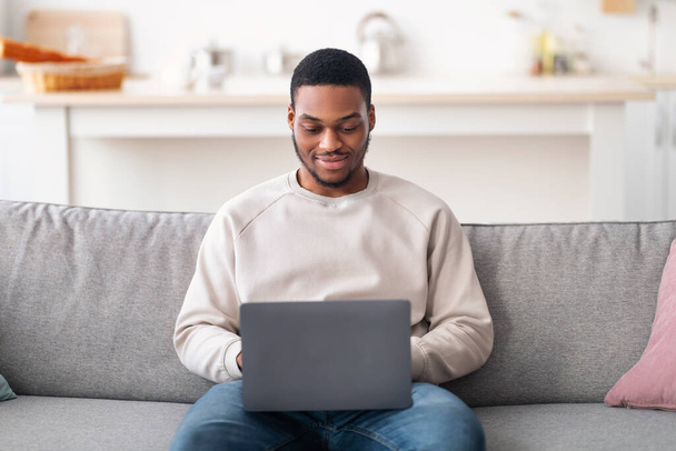 Sorridente ragazzo nero utilizzando il computer portatile, seduto sul divano - Foto, immagini