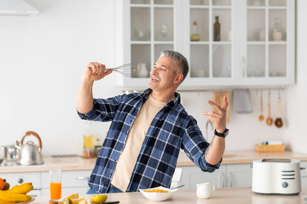 Hombre mayor engañando y cantando en batidor mientras cocina el desayuno en el interior de la cocina moderna. - Foto, Imagen