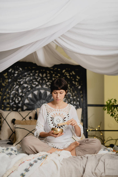 Young woman holding Tibetan singing bowl and pestle sitting on the bed. - Foto, afbeelding