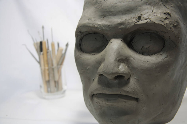 Мужское лицо в глине с инструментами для скульптуры. Творчество в процессе создания, на белом фоне - Фото, изображение